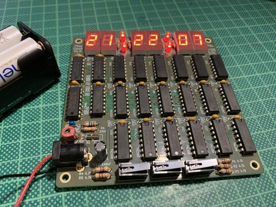 A square, green PCB on a green cutting mat. The PCB has three rows of TTL logic chips. At the top six seven segment LED displays are visible showing a time of 21:22:07.