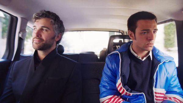 A still from the film Duino. A happy, serene-looking forty-something Argentinean man and a miserable looking Argentinean teenager sit side by side in a minibus as it drives along a leafy street.