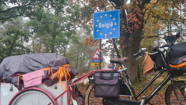 The bike and wagon are standing in front of a sign that says Belgium. It's a blue sign with white text which is surrounded by a circle of yellow stars.