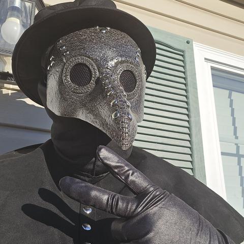 Selfie of a person dressed in an all black plague doctor costume with a small top hat giving a peace sign in front of a green shudder / window. A porch light can be seen from behind their head.