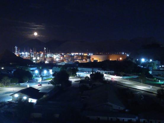 A photo of a city at night. The sky is dark and the moon is full and low. Some light clouds can be seen around the moon with one of them slightly covering it's bottom part. Under the moon, the silhouette of some mountains can be barely appreciated. In front of the mountain there are the lights of a refinery and a road in front of the refinery. On this side of the road there are the lights of a small one-floor house.