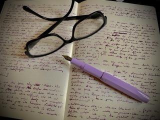 Verkleinertes und unscharfes Foto eines Schreibbuchs, dicht inkleiner Schrift beschrieben. Darauf liegt ein fliederfarbener Kaweco-Füller und eine schwarze Lesebrille