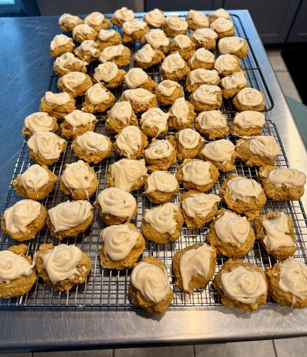 A wire cooling rack filled with numerous small, round cookies topped with swirls of frosting. The cookies have a golden-brown color, and the frosting is light in color, creating a visually appealing dessert display.
