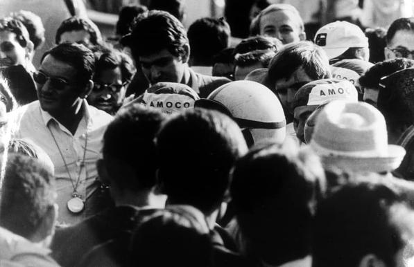 A black and white photo depicting a bustling crowd, with many individuals donning baseball caps emblazoned with "AMOCO" logos. A central figure appears to be surrounded by the gathered people, some of whom are wearing sunglasses or holding items like hats and cameras, suggesting an outdoor event during daylight hours.