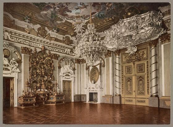 The image depicts an ornate and grandiose interior, likely a throne room or hall within a royal palace. The ceiling is adorned with elaborate frescoes depicting celestial scenes with clouds and mythological figures. A large chandelier hangs prominently from the center of the vaulted ceiling, adding to the opulence.
Below, on the walls and columns are intricate gold embellishments, including gilded decorations that resemble floral motifs and classical friezes. Large, ornate doors frame one side of the room, with golden panels featuring detailed engravings or carvings. The furniture includes a throne-like chair with elaborate canopy work above it.
The flooring is composed of large wooden planks, while to the left stands an impressive fireplace mantel embellished with gold and mirrors reflecting parts of the room's interior. Overall, the space exudes regal splendor through its use of rich colors, luxurious materials, and sophisticated design elements characteristic of European royal architecture during a historical period known for such opulence.