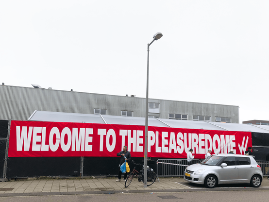 Large red banner with white letters stretched across a fence with balck tarps. The banner reads WELCOME TO THE PLEASUREDOME. On the pavement in front of the banner stands a man holding a blue and a yellow bag. To the right in the photo is a small silver car parked on the street. In the middle of the photo is a tall lamp post.