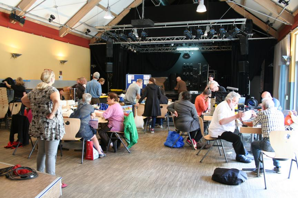 Eine Reparaturveranstaltung in einem Saal: 18 Personen stehen und sitzen um Tische verteilt, an denen mit verschiedenen Gewerken (Löten, Nähen usw.) gemeinsam repariert wird
