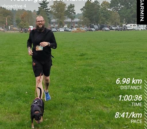 Ein Mann in kurzer schwarzer Laufbekleidung läuft mit einem Hund im Zuggeschirr über eine grüne Wiese. 
Das Zuggeschirr ist mit einem Leine an einem Hüftgurt befestigt, der schwer zu erkennen ist, da alles wie die Bekleidung gleich schwarz ist.
Im Hintergrund der Parkplatz des Veranstaltungsgeländes. 

Rechts unten im Bild sind die Daten des Laufs angegeben. 
6,98km Distance. 1 Stunde, 36 Sekunden.
8 Minuten 41 Sekunden pro Kilometer Pace.