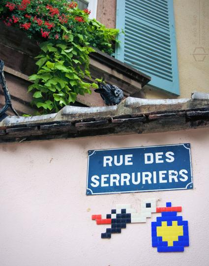 Picture of a wall with the street sign "Rue des Serrurier" and an invader tile art of an stork.