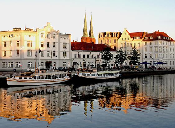 Vorn Flusswasser. In der Mitte zwei am Ufer liegende Schiffe. Darüber Häuser und zwei Kirchtürme, die sich auch vorn im Wasser spiegeln.
