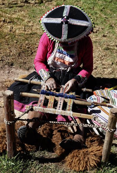 A person dressed in traditional clothing is weaving on a wooden loom outdoors. The individual is wearing a large, decorative hat and is focused on the weaving process. Surrounding them are natural elements, including grass and fabric pieces.