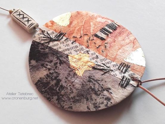 A round pendant with 2 different rust prints for each half: a nearly black one from plants and a rusty brown one with pure rust. A piece of an old book page separates the halves. Abstract embroidery with finest paper yarn, guilding, and hand-carved bone beds.