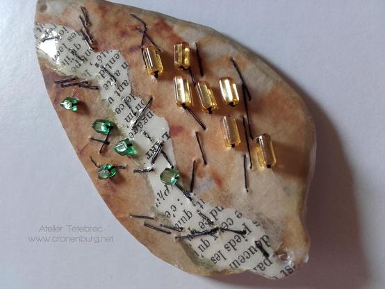 An autumn beech leaf made with paper art: rust print and parts of an old book page. Embroidery with the finest black paper yarn and sewn-on Preciosa crystal glass beads in yellow and green.