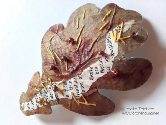 A paper art oak leaf made with rust print and parts of an old book page. Some stitches with yellow yearn and sewn-on Picasso beads from a glass manufactory.