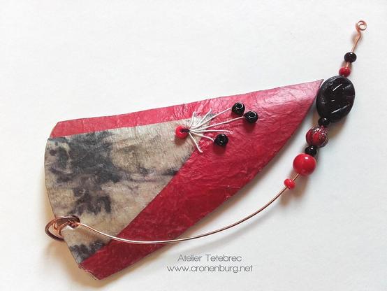 Abstract modern style of a paper art pendant. A blackish rust print of leaves is applied on red, hand-made mulberry paper. Radiant embroidery with white paper yarn and a red and 3 black seed beads. A collection of black and red glass artist's beads from a manufactory sits on a bent wire underneath.