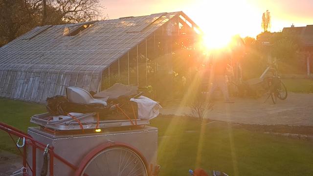 The man from before is in his backyard, the sun breaking into the view, shining over him, as he walks out of his greenhouse. Cart in the foreground.