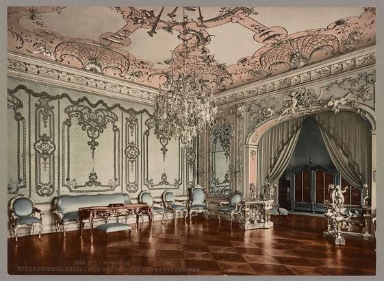 The image displays a luxurious bedroom with elaborate Baroque-style decoration. The room has polished wooden floors and high ceilings adorned with intricate plasterwork, including decorative moldings, scrolls, and garlands that add to its opulent appearance.
At the center of the space is an ornate chandelier hanging from the ceiling, contributing to the grandeur. Two large windows dressed in heavy curtains allow a modest amount of light into the room while maintaining privacy behind them. A fireplace with a mantel acts as a focal point and adds warmth to the otherwise cold color palette dominated by muted tones.
Furniture includes several upholstered chairs and ornate tables, reflecting an era where comfort was paired with grandeur in design. The walls feature large, patterned wallpaper that complements the room's lavishness. Overall, this setting suggests a residence belonging to someone of significant wealth or status during the 18th century European aristocracy.