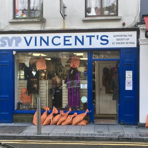 Blaue Fensterfront. Im Schaufenster orangefarbene Halloween-Deko. Vor dem Laden eine Reihe orangefarbener Sandsäcke.
Blue shop window front. Orange Halloween decorations in the shop window. A row of orange-coloured sandbags in front of the shop.