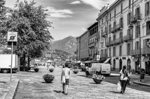 A pedestrian street scene in black and white featuring a man walking toward the camera and a woman carrying shopping bags. The background includes buildings, decorative flower pots, and trees, with mountains visible in the distance. A sign indicates a pedestrian area.