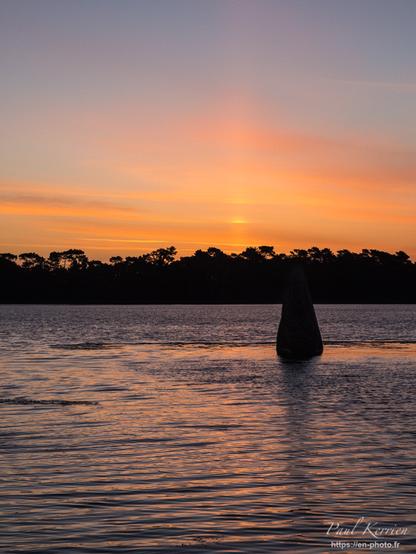 Photo en couleur en format vertical, présentant dans la moitié inférieure du cadrage un menhir se reflétant dans l'eau de la pleine mer qui le ceinture et dans la moitié supérieure du cadrage, des arbres de la rive opposée, et au-dessus le ciel de l'aube avec notamment un pilier solaire, bien vertical dans le ciel partiellement ennuagé. Evidemment, que ce soit les nuages, dans le ciel ou la surface de la mer sont colorés en orange / rose.