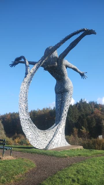 View from behind the tall, steel statue of the mermaid Arria, arms outstretched, the curve of her tail elegantly swooping around, lines of autumn trees in the foreground