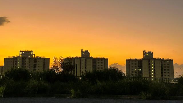 Silhouette of three apartment buildings against a vibrant orange and yellow sunset, with grass and plants in the foreground.