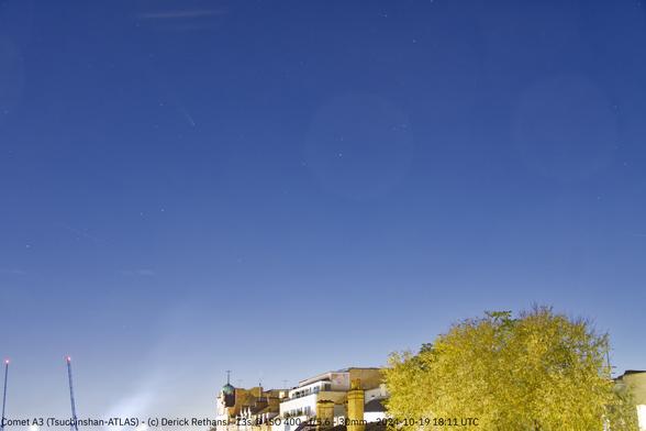 A blueish sky with two cranes on the horizon on the left, the top of buildings in the middle, and some trees on the right. In the sky, about a third from the left and a quarter from the top is a comet.