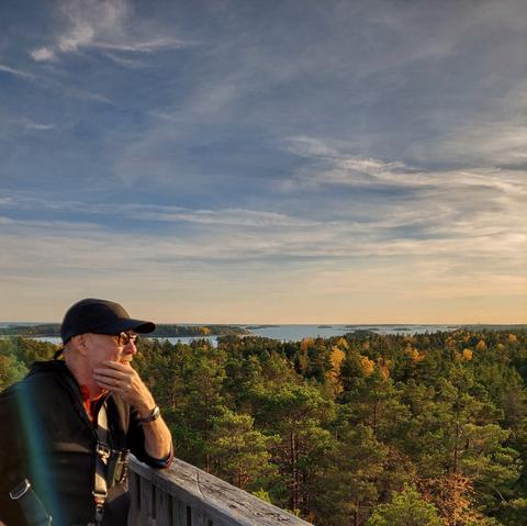 A man with his hand on his chin, wearing black baseball cap and clothes, stands looking over the wooden railing on top of a tower surrounded by forest in autumn foliage and sea in the distance
