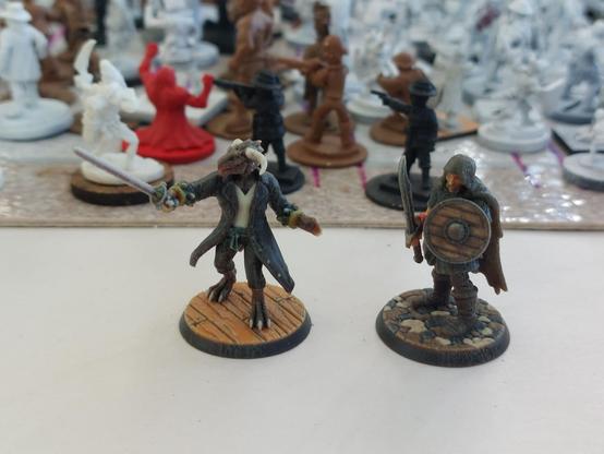 Two Hero Forge color printed miniatures: a dragonborn swashbuckler and a hooded human warrior with a falchion.