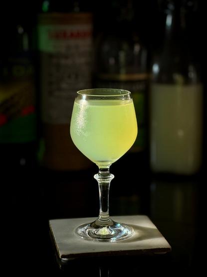 bright green cocktail in a chilled coupe glass sitting on a white coaster in the sunlight with all the bottles of ingredients behind it