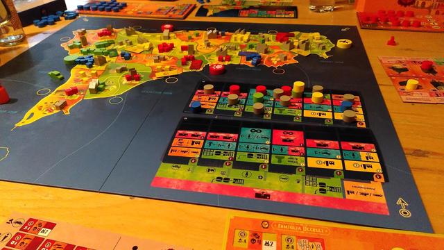 Aufgebautes Brettspiel La Famiglia nach dem Ende der ersten Planungsphase. Im Vordergrund zwei Aktionstableaus mit verschiedenen Steinen auf verschiedenen Aktionsfeldern. Im Hintergrund der Spielplan mit eingesetzen soldaten (holzwürfel) autos und drogenlaboren.