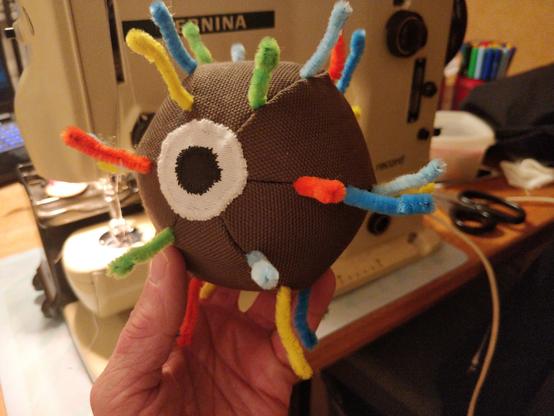 A hand holding a brown-green cloth ball with colourful arms (made from pipe cleaners) sticking out in all directions. A single large eye is looking to the left. In the background there is an old Bernina sewing machine and sewing supplies.