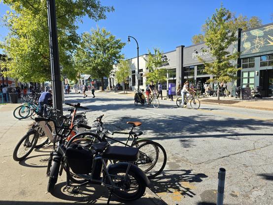 People walk and bike on a street closed to cars on a sunny day. Many bikes are parked along the sidewalk.