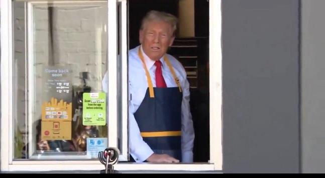 The Dear Leader poses in the drive-up window of a McDonalds restaurant. He's wearing a blue server's apron and looks extremely uncomfortable.