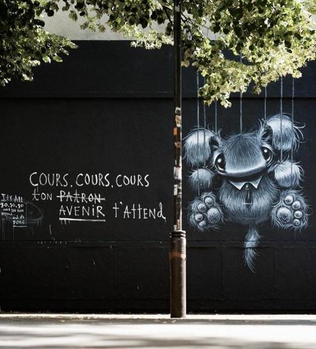 Streetartwall. A fluffy fantasy creature with a message was sprayed/painted on a black painted wall with changing street art. A small, gray, fluffy creature with very large eyes, curved ears and a broad nose hangs on thin ropes on the wall and looks at us. Next to it is the text in French: Run, run, run, your (boss's) future awaits. The word boss is crossed out.