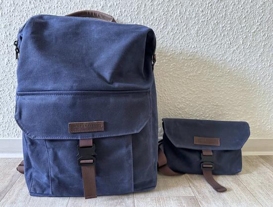 Dunkelblauer Waterfield Vitesse Backpack und kleine, ebenso dunkelblaue Umhängetasche Vitesse Sling, stehen nebeneinander auf dem Boden vor einer weißen Wand.