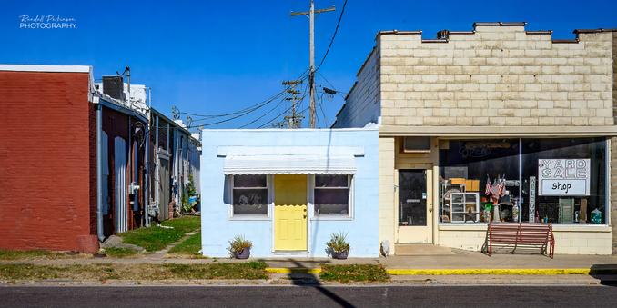 A very small building with a yellow front door and two windows under an awning sits next to other commercial buildings on a street in a small town.