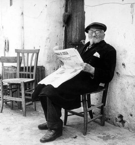 A black and white photograph of an elderly man seated on a wooden chair, leaning against a weathered wall. He is dressed in dark clothing with a coat, trousers, shoes, and a hat complementing his attire. His right hand holds open a newspaper titled "L'Unità", which appears to be from the year 1963 based on its layout and style of print. The man has glasses perched atop his nose, a white beard, and is looking directly at the camera with an attentive expression. Surrounding him are several other wooden chairs, suggesting that he might be in a public or communal space such as a café or park.