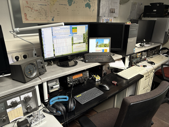 A fixed amateur radio station setup with a rotor controller, two monitors, headphones, a transceiver and morse key