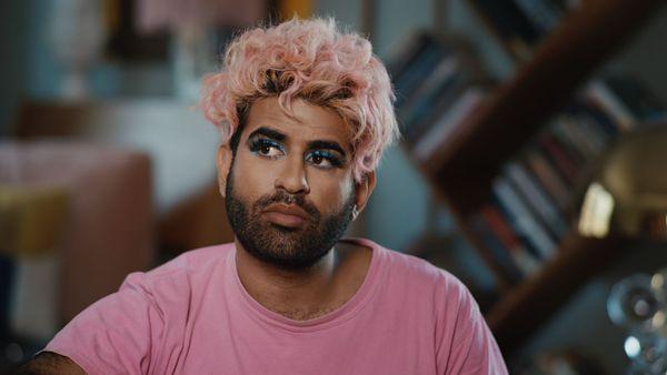 A still from the film Alok. A non-binary person with light brown skin, curly pink hair, short black facial hair, blue eyeshadow and a pink t-shirt sits in front of tilted bookshelves and a pile of cardboard boxes.