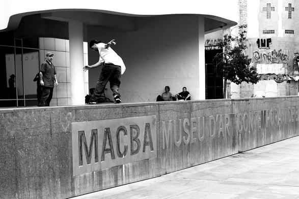 A skateboarder doing a backside tailslide on a stone ledge. The wall below the ledge is embossed with the word MACBA