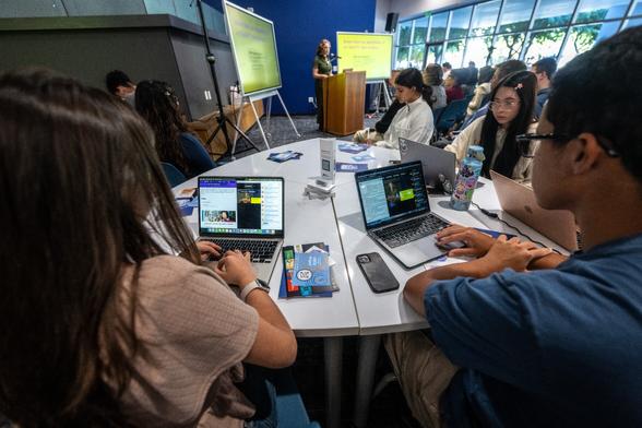 students on laptops helping the online community connect to Lauren Klein's talk with 2 screens with slides facing the audience