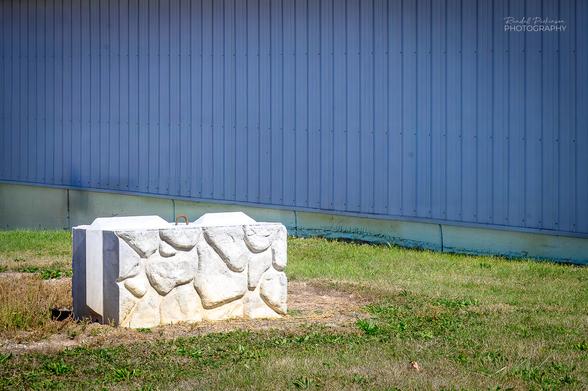 a single large retaining wall concrete block with a stone patterned face sits in a yard next to a blue metal building.