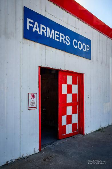 A white building with bright red trim, a double door painted in a red and white checkerboard pattern, and a blue sign over the door reading "FARMERS COOP" in white letters.