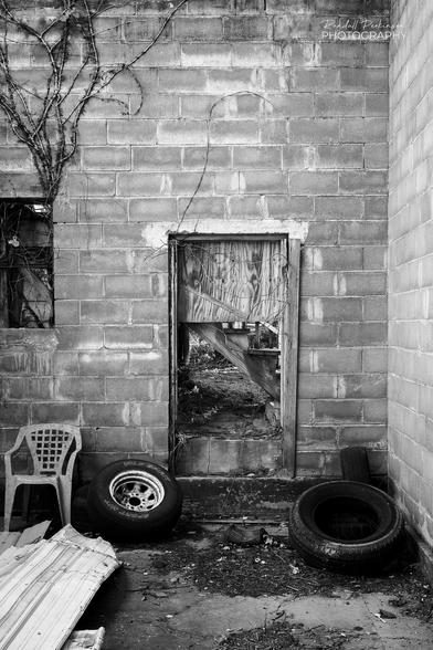 Concrete block walls of a building which no longer has a roof.  Two old tires, a plastic chair sit in the room.  A wooden door in the back wall has a big square hole in it.  Ivy is growing up the wall from a window opening.