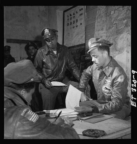 The image is a black and white photograph depicting four individuals, presumably military personnel from the 332nd Fighter Group during World War II. The primary focus of the photo shows two men seated at a desk, with one man standing behind them.
One seated individual wears an aviator's cap adorned with several insignias, indicating his rank or affiliation within the group. He is dressed in a leather flight jacket and appears to be engaged in reviewing documents on the table before him, which includes what seems like maps or charts related to military operations.
The other man at the desk, who is also standing but slightly turned away from us, wears similar attire with additional patches indicating his role as an officer. He holds another document and looks attentively towards something outside of this frame.

Behind these two individuals are more men seated in a semi-circle arrangement around what appears to be a table or a planning surface. The background is cluttered with various items such as maps, charts, and other military paraphernalia that suggest strategic discussions were taking place.
The overall atmosphere conveys the seriousness and intensity of wartime operations being discussed among these individuals.