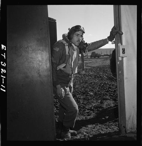 The image is a black and white photograph of an individual identified as Edward M. Thomas, a Tuskegee airman from Chicago, IL who was part of Class 43-J stationed at Ramitelli in Italy during March 1945. The person stands next to what appears to be the entrance or doorway of a military structure with rugged terrain visible behind them.
Edward is wearing an aviator's helmet and goggles attached to his flight suit, which includes patches indicating rank and unit affiliation. He holds onto a handle on the doorframe while his left hand hangs by his side. The environment suggests he might have been flying missions during World War II as part of the famous Tuskegee Airmen squadron.
The photograph is labeled with "T23-1" in text, which could be an image reference or cataloging code used for archival purposes.