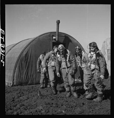 The image depicts a group of four individuals in military uniforms, likely soldiers or pilots during World War II. They are standing on what appears to be muddy ground and walking away from an oversized canvas tent-like structure with a prominent chimney pipe sticking out the top.

Each person is dressed similarly: wearing heavy coats, gloves, boots, and carrying equipment such as helmets and what seems like parachutes or other gear strapped around their bodies. The uniforms are indicative of military attire during that era, possibly for cold-weather operations given the thick clothing material visible in some sections.

The men exhibit focused expressions on their faces while looking ahead, suggesting a sense of purpose or determination. Their poses indicate movement as they stride forward together out of frame towards another destination outside this captured moment.

In summary, the image captures Tuskegee Airmen exiting what appears to be a parachute room located in Ramitelli, Italy during March 1945. The photograph is black and white with visible wear around its edges indicating age or handling over time.