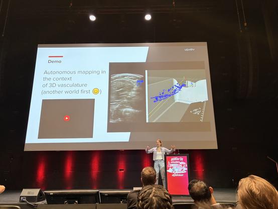 Deanna stands on stage in front of a huge screen. Her hands are gesturing openly. A demo video of real time vascular mapping plays on the screen behind her.