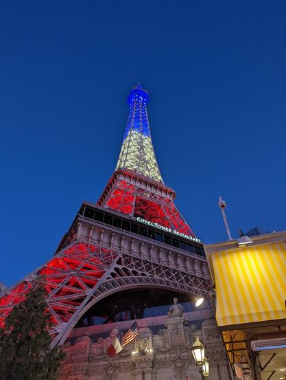 A photo I took of the Eiffel Tower that's at the Paris Las Vegas Hotel and Casino in Las Vegas. You can see the tower is lit up in red, white and blue, and you can see various structures around it as well as the night time sky behind it.